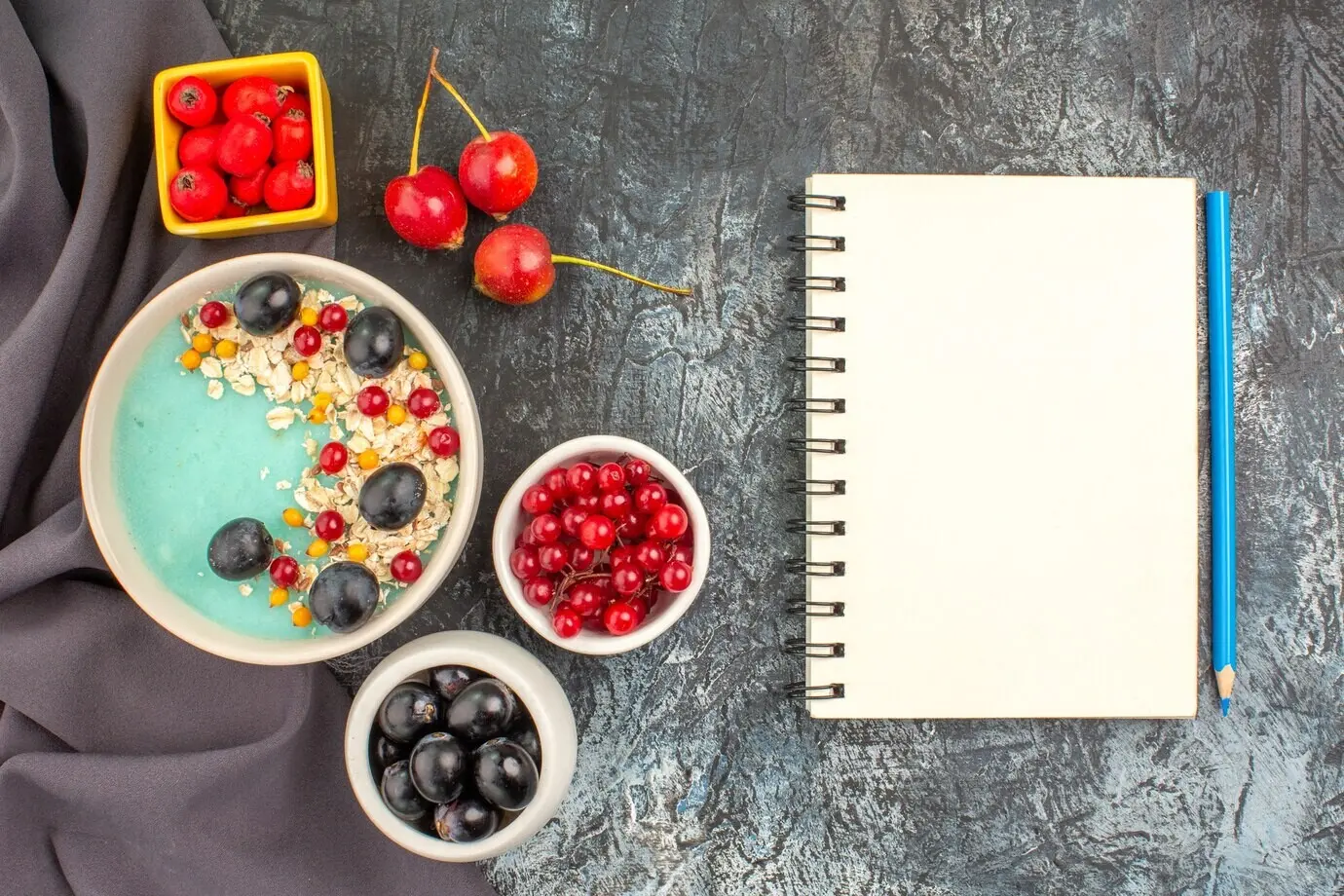 Nahaufnahme aus der Draufsicht von Haferbrei mit Beeren, bunten Beeren, einem Granatapfel, einem Notizbuch und einem Bleistift auf der Tischdecke.