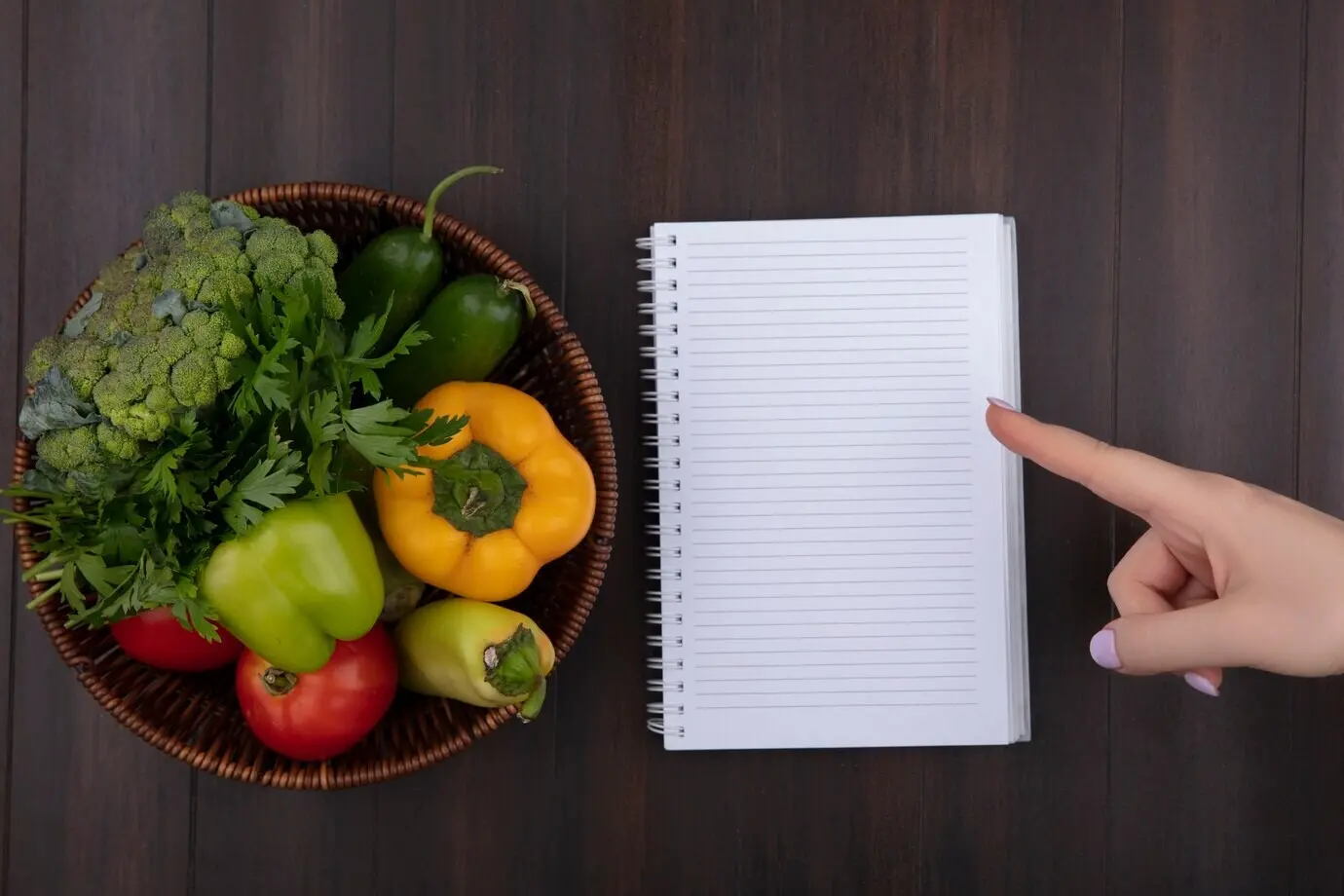 Draufsicht mit Copyspace: Eine Frau zeigt auf ein Notizbuch; Paprika, Petersilie und Tomaten in einem Korb auf Holzhintergrund.